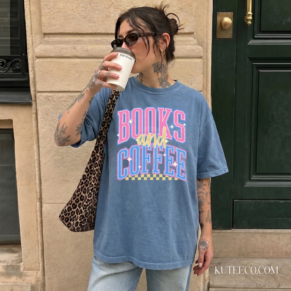 A woman wearing an oversized blue graphic t-shirt with 'BOOKS AND COFFEE' printed in pink, yellow, and blue retro lettering. She is holding a white paper coffee cup and wearing a leopard-print shoulder bag, standing in front of a beige stone wall and a dark green door.