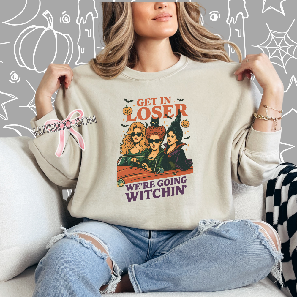 Person wearing a beige sweatshirt with 'Hocus Pocus' design, sitting on a couch with Halloween-themed background. hocus pocus get in loser were going witchin sweatshirt 